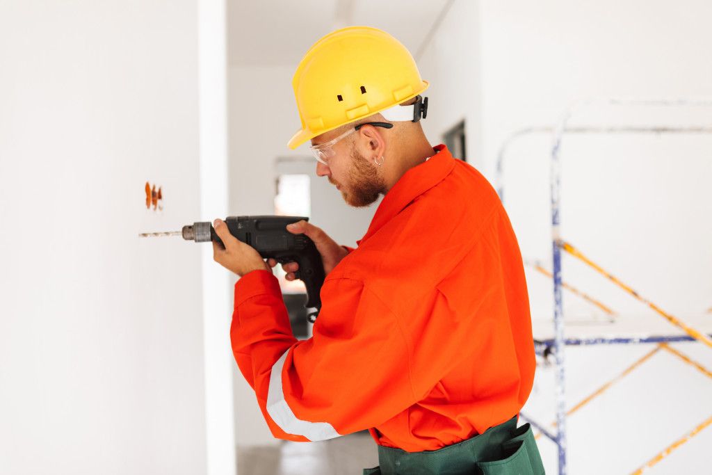 young-foreman-in-orange-work-clothes-and-yellow-hardhat-using-electric-drill-at-work-in-new-apartments.jpg
